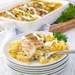 A plated serving of baked chicken with rice, garnished with sauce and parsley, is in focus. In the background, a casserole dish holds the remaining chicken and rice. The setting includes a white and blue striped towel and fresh herbs.