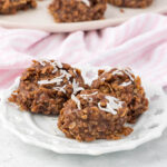 Three chocolate oatmeal cookies topped with shredded coconut are placed on a decorative white plate. A pink and white striped cloth is partially visible in the background.