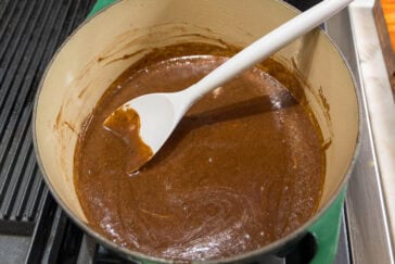 A thick, dark brown sauce is being stirred in a green pot on a stove. A white spatula is resting inside the pot, partially covered in the sauce. The stove's black grates are visible on the left side.