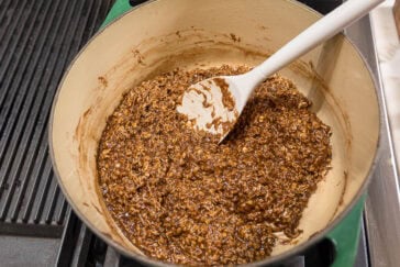 A pot filled with a mixture of chocolate, oats, and other ingredients on a stovetop. A white spatula is resting in the mixture, ready for stirring. The pot is placed on a black grill-like stove surface.