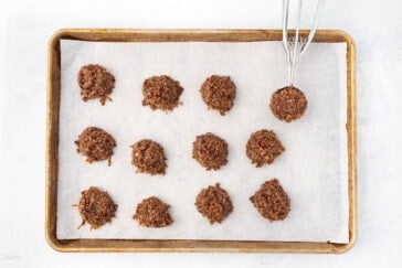A baking sheet lined with parchment paper holds twelve scoops of oatmeal chocolate cookie dough. A metal cookie scoop with dough is placed in the upper right corner, suggesting more cookies to be made.