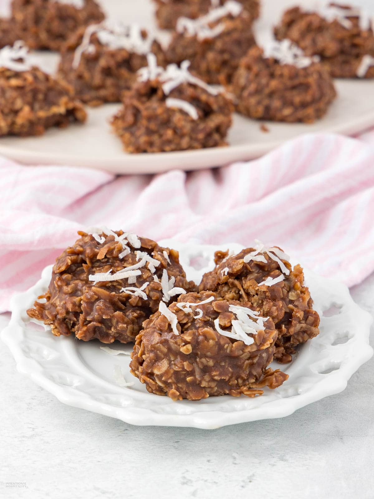 Three no-bake chocolate oatmeal cookies topped with shredded coconut on a white plate. More cookies are in the background on a pink-striped cloth.