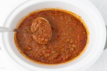 A white slow cooker filled with hearty chili, featuring ground meat, tomatoes, and beans, being stirred with a ladle. The chili appears thick and savory, with a rich reddish color.