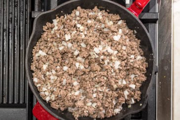 A cast iron skillet on a stovetop contains cooked ground beef mixed with chopped onions. The skillet handle is red, and the stovetop has a black grill surface next to it.