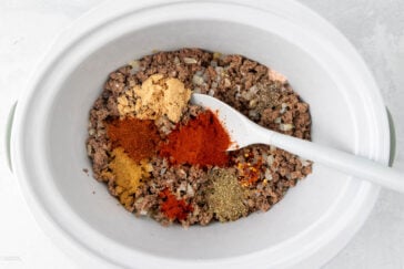 Ground meat and chopped onions in a slow cooker, topped with various vibrant spices including paprika, chili powder, and cumin. A white spoon rests in the mixture, ready for stirring.