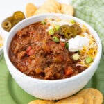 A bowl of chili topped with shredded cheese, sliced jalapeños, sour cream, and chopped green onions. The bowl is surrounded by crackers and sliced jalapeños on a green plate. A green cloth napkin is in the background.
