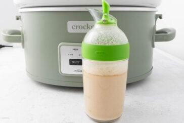 A green Crockpot with a control panel displaying cooking time settings is in the background. In the foreground, there is a clear Oxo container with a green lid, filled with a frothy beige liquid.