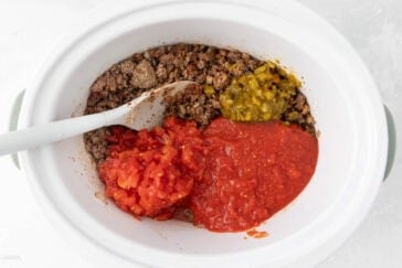 A white crockpot with cooked ground beef, chopped tomatoes, tomato sauce, and diced green peppers, being mixed with a white spoon.