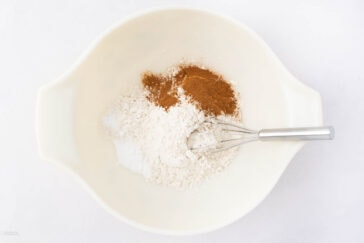 A white mixing bowl contains flour, cinnamon, and baking soda. A whisk rests in the bowl, set against a plain white background.