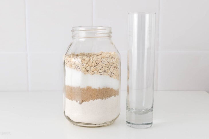 A glass jar filled with layered ingredients, including oats, sugar, brown sugar, and flour, sits next to a tall empty glass on a white countertop against a tiled white background.