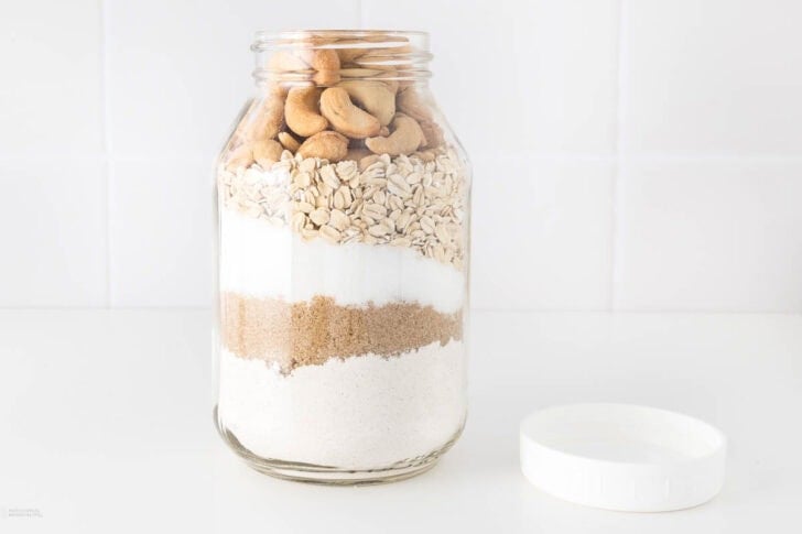 A glass jar filled with layers of cookie ingredients, including flour, brown sugar, white sugar, oats, and topped with cookies. The jar is on a white surface with a white lid beside it, set against a white tiled background.