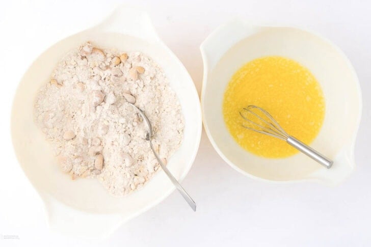 Two white bowls on a countertop: the left bowl contains dry ingredients with a spoon for mixing, and the right bowl contains melted butter with a whisk.