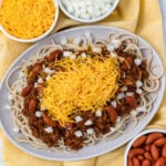 A plate of spaghetti topped with chili, red kidney beans, chopped onions, and shredded cheddar cheese. Surrounding the plate are bowls of extra cheddar cheese, diced onions, and kidney beans. A yellow cloth is placed underneath.