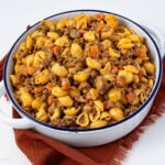 A dish filled with a hearty pasta meal consisting of shell pasta, ground meat, and diced tomatoes. The food is in a large, round white dish with handles, resting on a burnt orange cloth.