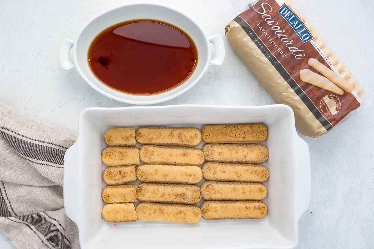 A white dish with a layer of soaked ladyfingers for tiramisu cake, a bowl of coffee, and an unopened package of ladyfingers on a light surface with a striped cloth nearby.