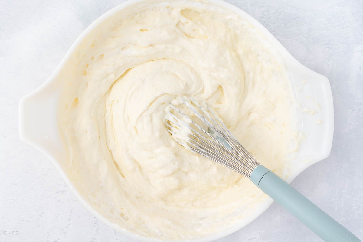 A whisk with a light blue handle rests in a white mixing bowl filled with creamy batter. The batter is smooth and has a pale yellow color, swirling around the utensil. The background is a light, textured surface.