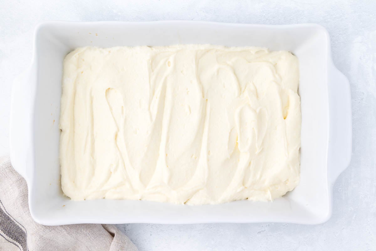 A white rectangular baking dish filled with smooth, creamy cheesecake batter. A striped cloth partially visible at the bottom left adds a cozy touch. The dish is set on a light gray textured surface.