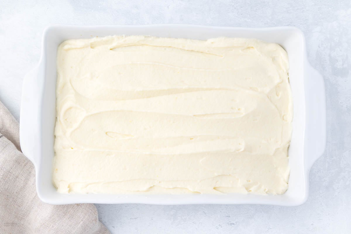 A rectangular white dish filled with smooth, creamy frosting, placed on a light-colored surface. A beige cloth is partially visible on the left side of the dish.