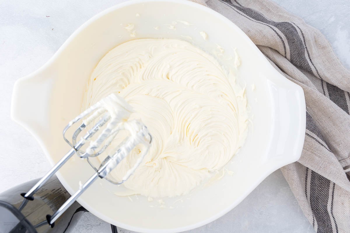 A white mixing bowl filled with creamy, whipped mixture for tiramisu cake sits on a light surface next to a striped kitchen towel; an electric hand mixer with beaters rests inside the bowl.