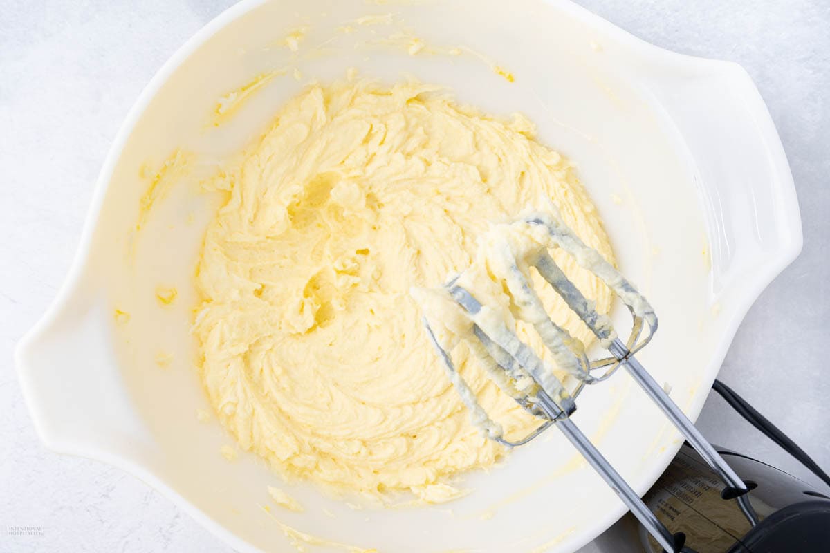 A white bowl filled with creamy, yellow cake batter. An electric hand mixer with batter-coated beaters rests on the rim. The background is a light, textured surface.