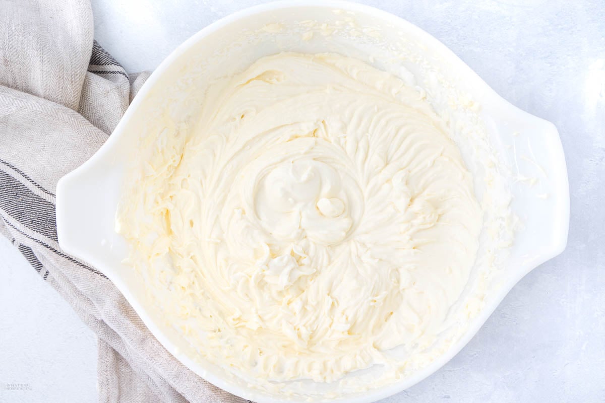 A white mixing bowl filled with creamy, smooth, whipped frosting. A striped cloth is partially visible beside the bowl on a light surface.