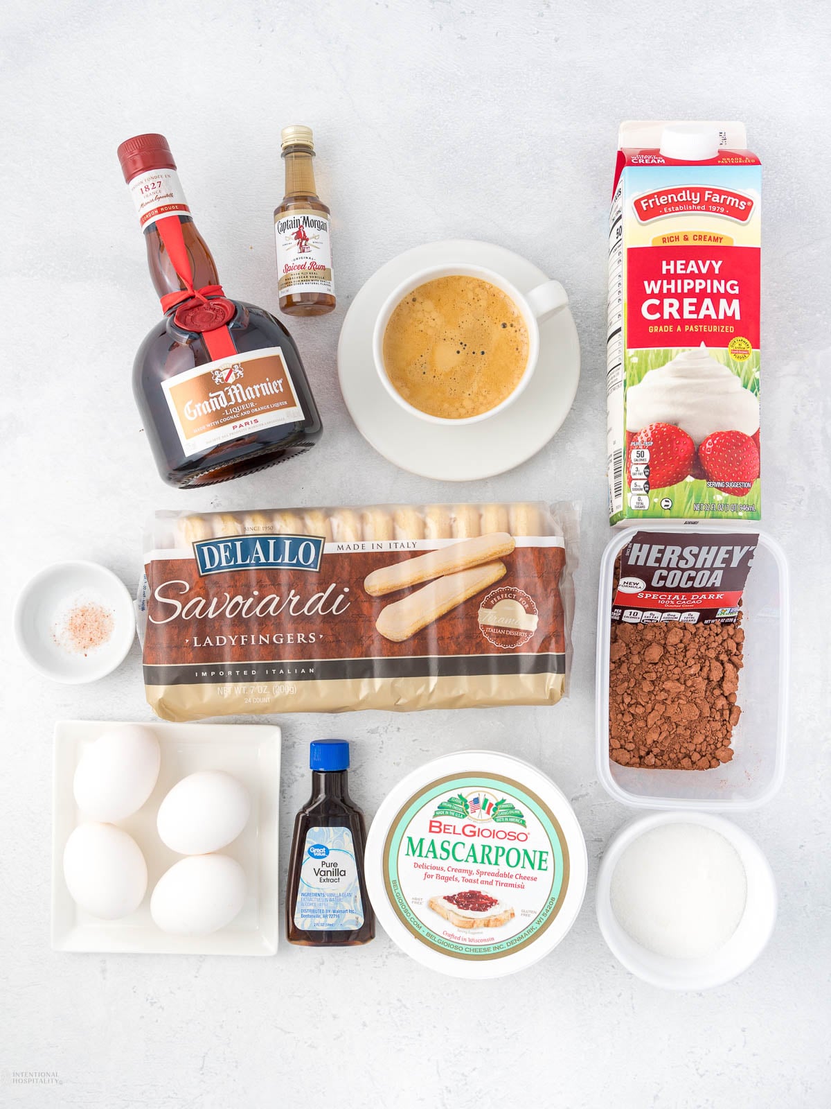 Ingredients for tiramisu laid out, including eggs, mascarpone, ladyfingers, cocoa powder, sugar, heavy cream, espresso, vanilla extract, Grand Marnier, coffee extract, and a pinch of salt, all arranged on a white surface.
