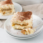 A slice of tiramisu on a stack of white plates, dusted with cocoa powder. A fork rests beside the dessert. A second plate with another slice is visible in the background, set on a light-colored tablecloth.