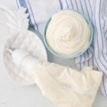 A bowl of whipped cream sits next to a piping bag filled with more cream on a white surface with a blue-striped towel.