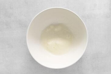 A white bowl with a smooth, fluffy mixture, likely whipped egg whites, against a light gray background.