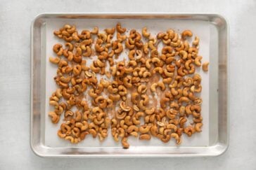 A metal baking tray filled with roasted cashews evenly spread out on a light gray surface. The cashews have a golden brown color, indicating they are toasted.
