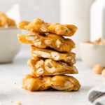 A stack of peanut brittle pieces with a golden-brown color, featuring visible peanuts embedded in the caramelized sugar. The background includes a blurred bowl and scattered peanuts on a light surface.