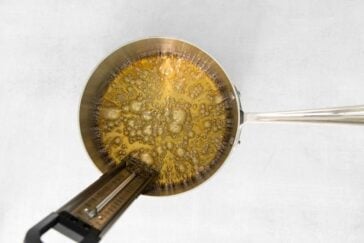 A pot on a stove with caramel bubbling inside. A metal candy thermometer is clipped to the side of the pot, measuring the temperature of the caramel. The background is a plain white surface.