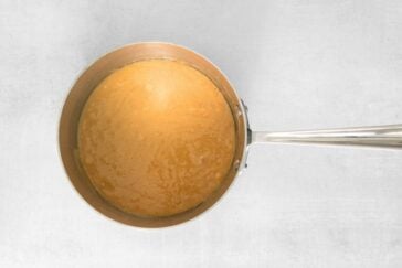 A silver saucepan filled with smooth caramel sauce on a light gray surface.