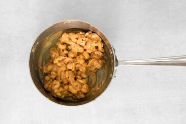 A metal saucepan filled with creamy mac and cheese is set on a light gray surface. The cheese sauce appears thick and well-mixed with the pasta. The pan has a long handle extending to the right.
