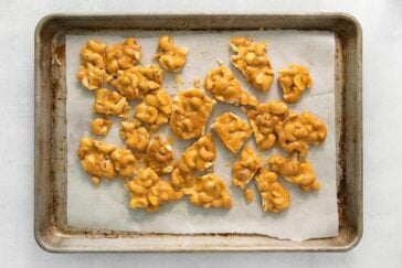A baking tray lined with parchment paper holds clusters of homemade cashew brittle. The golden, glossy candy is spread out, showcasing a mix of crunchy cashews embedded in hardened caramelized sugar.