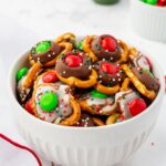 A white bowl filled with holiday-themed treats. The treats consist of pretzels topped with melted chocolate and colorful red and green candies, sprinkled with red, green, and white nonpareils. The background is softly blurred.