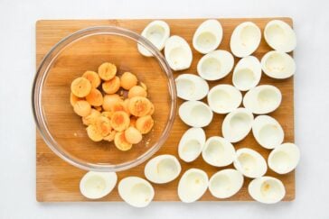 Halved hard-boiled eggs arranged on a wooden cutting board. The yolks are placed in a clear bowl on the board's left side.
