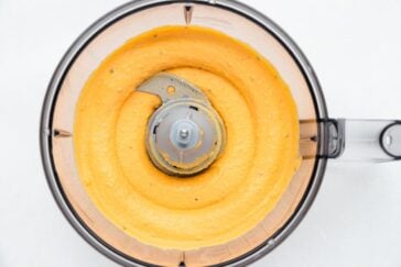 Top view of a food processor bowl containing smooth, creamy orange hummus. The hummus is evenly spread in a spiral pattern around the central blade. The food processor has a transparent plastic lid.