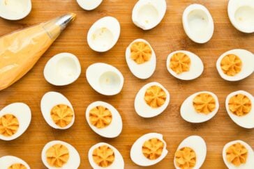Halved hard-boiled eggs with the yolks removed, ready to be filled with a creamy filling from a nearby piping bag, are arranged on a wooden surface. The filling is piped into several egg halves, creating a decorative pattern.