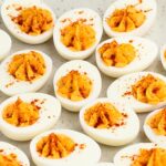 A platter of deviled eggs neatly arranged. Each half has a creamy filling topped with a sprinkle of paprika, giving a vibrant contrast to the white of the egg whites. The background is a speckled light gray surface.