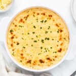 A baked dish of creamy, golden cheese dip garnished with chopped herbs in a round, white ceramic dish. The surface is lightly browned, giving it a crispy appearance. A small bowl of shredded cheese is visible in the top left corner.