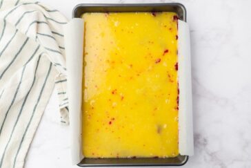 A rectangular baking pan filled with a bright yellow mixture, possibly cake batter, dotted with red specks. The pan is lined with parchment paper. A white and black striped kitchen towel is placed beside the pan on a marble surface.