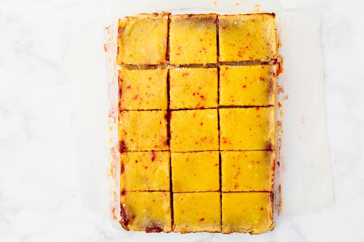 Top view of a rectangular tray of sliced lemon bars arranged in a grid. The bars have a golden yellow filling with a light dusting of powdered sugar on top and a thin crust at the bottom. They appear to be on white parchment paper.
