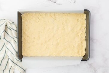 A rectangular baking pan lined with parchment paper is filled with an even layer of pale, unbaked dough. A striped kitchen towel is partially visible to the side on a marble surface.