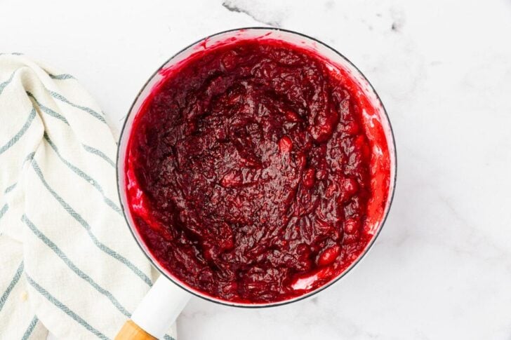 A pot of thick, rich cranberry sauce with a deep red color sits on a white marble surface. A light-colored cloth with dark stripes is partially visible beside the pot.