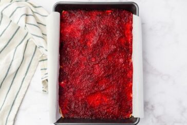 A rectangle baking dish with a red, berry-covered dessert on parchment paper. A striped cloth is visible on the left side on a marble countertop.