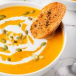 A bowl of creamy pumpkin soup garnished with a swirl of cream and sprinkled with pumpkin seeds. A slice of toasted bread rests on the edge of the bowl. The bowl is set on a white marble surface with a spoon beside it.
