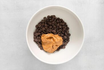 A white bowl containing a pile of chocolate chips with a dollop of peanut butter on top, placed on a light gray surface.