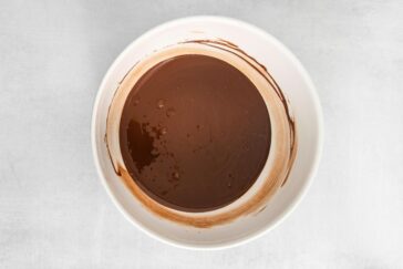 A white bowl containing a smooth, melted chocolate mixture, with some remnants smeared along the sides. The background is a light gray surface.