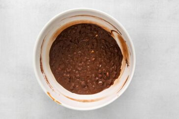 A bowl filled with chocolate and nuts mixture sits on a light gray surface. The mixture is thick and slightly textured, suggesting it might be a chocolate nut batter or filling.